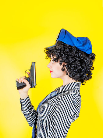 Funny woman in an American police cap and dress with a gun, on a yellow backgroundの写真素材