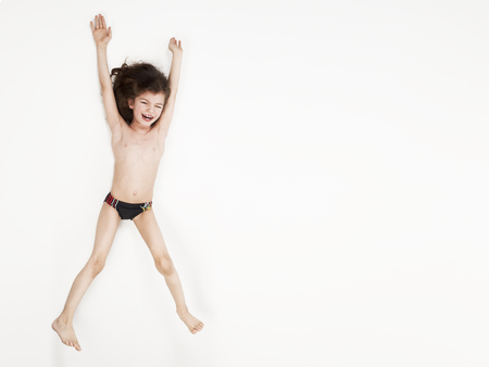 Smiling little boy in swimming trunks jumping with arms raised against a white backgroundの写真素材