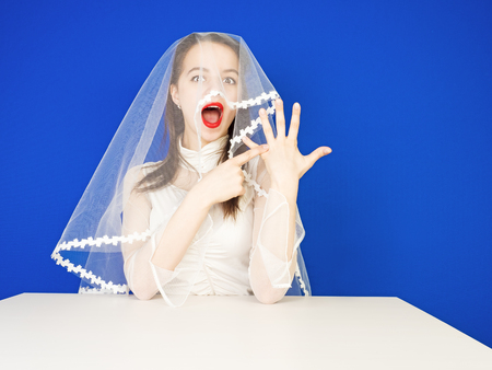 The happy bride boastfully and arrogantly shows the ring on her finger while sitting at the table. The blue backgroundの写真素材