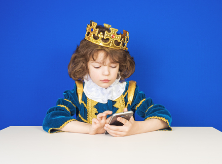 Cute little boy in a king costume using a modern smartphone on a blue background.の写真素材