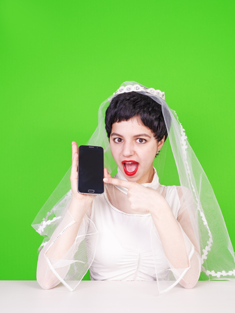 Caucasian fiancee holding a mobile phone and pointing at it. Young fiancee with a mobile phone. Fiancee using a mobile phone sitting at a table on a green background.の写真素材