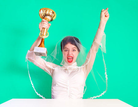 Caucasian bride in a wedding attire holding a golden trophy. Half length of young brunette with a trophy. Happy bride sitting at a table with a trophy on a turquoise background.の写真素材