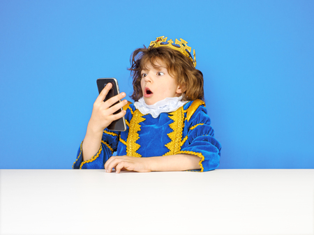 Portrait of a child in a king dress, surprised boy, little man using a smart phone on a colored background. Funny looking human face expression, emotion, reaction, body language. Communicationの写真素材
