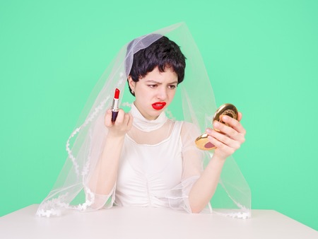 Young enraged bride teenager looking at her hairstyle in a Mirror sitting at table isolated on light backgroundの写真素材