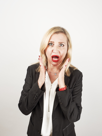 Blond female teacher wearing a strict suit keeping hands on head, terrified in panic, joking against a light backgroundの写真素材