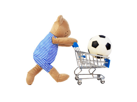 A toy bear carries a shopping trolley with a football. Sports goods and toys falling into the basket, isolated on white.の写真素材
