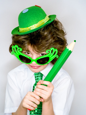 Funny St Patricks day stunning little boy wearing a green hat, carnival glasses and a tie. Curly child posing with a big green pencil in hand on a light backgroundの写真素材