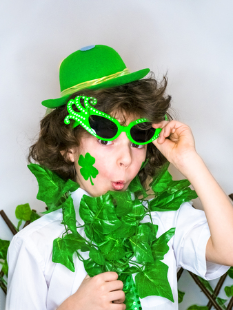 Funny St Patricks day little curly boy wearing a green hat and green carnival accessories shocked with an open mouth. light backgroundの写真素材