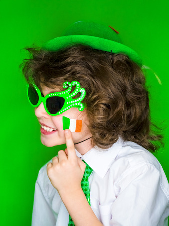 Child Celebrating St. Patrick's Day Showing his Make-up. A small, curly boy in green carnival accessories points his finger at Irish flag on his cheek. green backgroundの写真素材