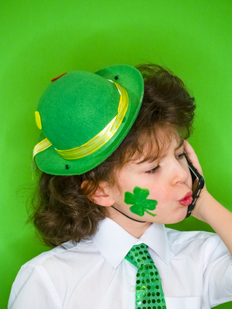 Funny St Patricks day stunning little boy wearing a green hat and shamrock, the Irish flag on his cheeks talking on the phone.Serious and thoughtful child listening to the news on the phoneの写真素材