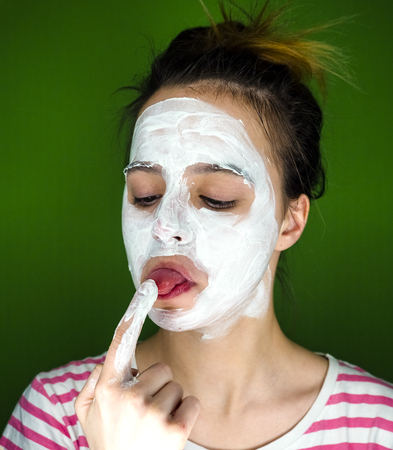 Close-up portrait of beautiful girl applying facial cream mask and beauty treatments on her face tasting her finger isolated onzelenom backgroundの写真素材