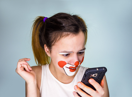 mime girl in white t-shirt with mobile phone in hand. Man with emotions on a white background. Surprised creepy, funny clown girl using smartphone isolated over grayの写真素材