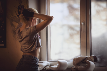 Blond boxer girl standing near the window strikes her hair before a fight. Portrait of a sportswoman in boxing belts in the gymの写真素材