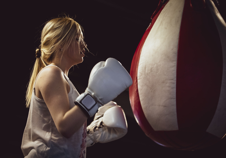 Portrait of sexy shaggy blonde boxer woman in white boxing gloves standing near red and white punching bagの写真素材