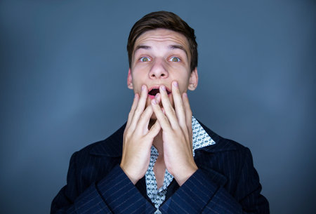 Horrified young man man covering mouth with hands looking at camera head shot portrait isolated on...
