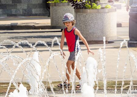 Child playing with water at street fountain in summer. Little boy playing in a fountain. excited boy running between water flow in city parkの写真素材