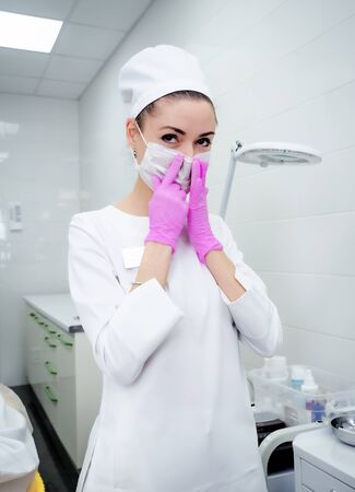 Young smiling brunette woman doctor, nurse, cosmetologist, chemist in stylish uniform and medical gloves is taking on holding medical mask on backgroundの写真素材