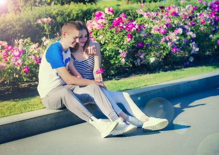 Loving couple sitting near the flowerbed with roses. Attractive woman and boyfriend in park, bush of roses flowers in the background. romantic date. man with girl in park. happy valentines day.の写真素材