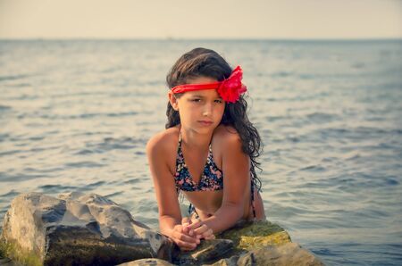 Mysterious dreamy young girl with a red bandage on her head sitting in the water on a rocky beachの写真素材