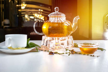 Fragrant fresh herbal tea and melissa in a glass teapot warming on a candle on a white table. Traditional herbal drink. Tea with honey. Kettle warmerの写真素材