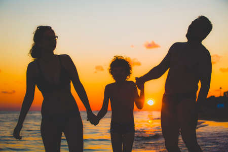 Dark silhouette of a happy family on a sunset.Father, mother, baby son walking along the sandy beach.Family having fun on the beach.Travel lifestyle, parents, dad with children on summer vacation.の写真素材