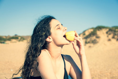 Brunette teen girl in a black swimsuit sitting by the sea, eating yellow pepper, healthy snack and...