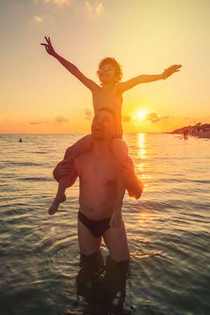 Black silhouettes of father and sana against the sunset sky. Mature father and son playing on beach. Curly Boy rising up hands imitating a flight at bright sunset through the shining sun.の写真素材