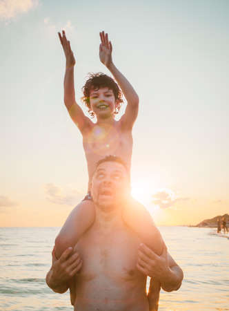Black silhouettes of father and sana against the sunset sky. Mature father and son playing on beach. Curly Boy rising up hands imitating a flight at bright sunset through the shining sun.の写真素材