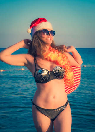 Beautiful young woman in Santa Hut, Hawaii accessory and bikini on beach. Christmas vacationPretty middle aged woman in black shiny swimsuit enjoying sand tropical beachの写真素材