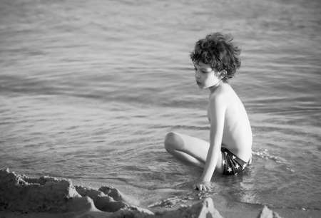 Cute curly male kid playing game at the beach.Little boy play with sand on summer beach.Funny games during summer vacation. Child having fun at coast. Leisure, games and vacation.Black and white photoの写真素材