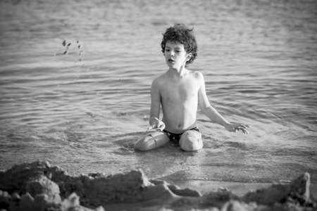 Cute curly male kid playing game at the beach.Little boy play with sand on summer beach.Funny games during summer vacation. Child having fun at coast. Leisure, games and vacation.Black and white photoの写真素材