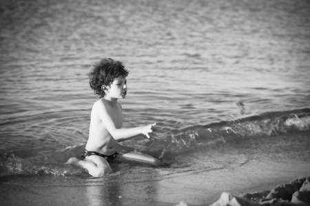 Cute curly male kid playing game at the beach.Little boy play with sand on summer beach.Funny games during summer vacation. Child having fun at coast. Leisure, games and vacation.Black and white photoの写真素材