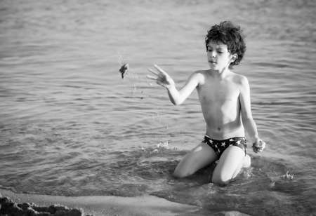 Cute curly male kid playing game at the beach.Little boy play with sand on summer beach.Funny games during summer vacation. Child having fun at coast. Leisure, games and vacation.Black and white photoの写真素材