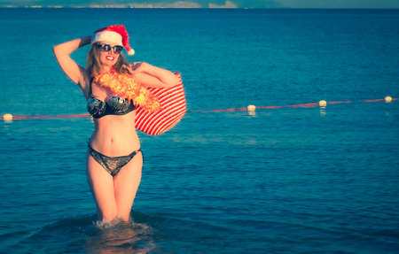 Beautiful young woman in Santa Hut, Hawaii accessory and bikini on beach. Christmas vacationPretty middle aged woman in black shiny swimsuit enjoying sand tropical beach. Copy spaceの写真素材
