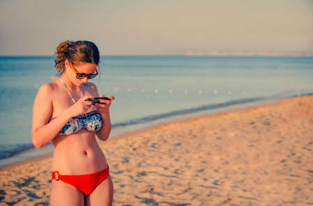 Pretty teenager girl brunette using a smart phone standing on the beach with the sea and horizon in the background. Copy spaceの写真素材