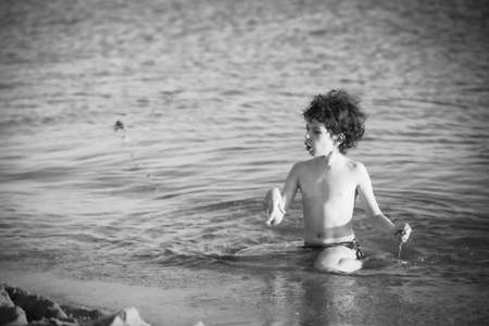 Cute curly male kid playing game at the beach.Little boy play with sand on summer beach.Funny games during summer vacation. Child having fun at coast. Leisure, games and vacation.Black and white photoの写真素材