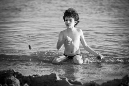 Cute curly male kid playing game at the beach.Little boy play with sand on summer beach.Funny games during summer vacation. Child having fun at coast. Leisure, games and vacation.Black and white photoの写真素材