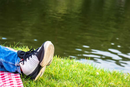 Woman's legs in sneakers and jeans resting on the lake shore. Female feet on green grass close up.の写真素材
