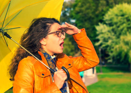 Shocked stylish young girl in leather jacket and with a yellow umbrella looking into the distance in the autumn Park. Autumn time. Copy spaceの写真素材