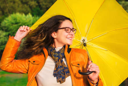 A smiling girl with a yellow umbrella is hiding from the rain.Beautiful brunette woman with glasses with umbrella on a rainy day.Attractive young woman carrying umbrella while standing in the parkの写真素材