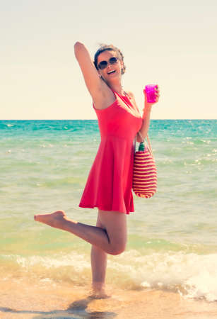 a woman brunette in a hat standing in the water on a sandy beach with a plastic glass in her hand on a clear summer day. vacation on sunny dayの写真素材