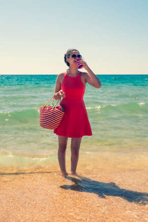 a woman brunette in a hat standing in the water on a sandy beach with a plastic glass in her hand on a clear summer day. vacation on sunny dayの写真素材