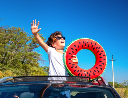 A little boy leaning out of the hatch of a car holding an inflatable ring is enjoying the journey and the trip to the sea. Happy child on the roof of the car raising his hands upの写真素材