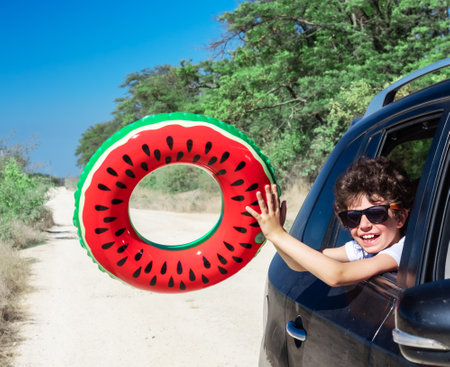 Family trip concept on the road to the sea: Portrait of two happy children swinging an inflatable ring at the camera, pouring their heads out of the car windowの写真素材
