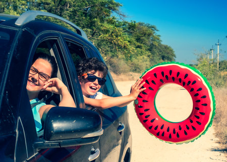 Family trip concept on the road to the sea: Portrait of two happy children swinging an inflatable ring at the camera, pouring their heads out of the car windowの写真素材