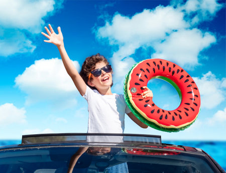 A little boy leaning out of the hatch of a car holding an inflatable ring is enjoying the journey and the trip to the sea. Happy child on the roof of the car raising his hands upの写真素材