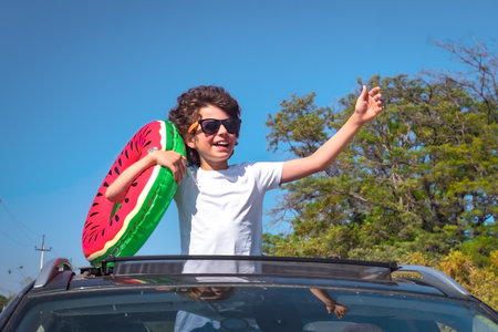 A little boy leaning out of the hatch of a car holding an inflatable ring is enjoying the journey and the trip to the sea. Happy child on the roof of the car raising his hands upの写真素材