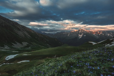 mountain peak sunset landscape with gloomy dramatic mainly cloudy sky and orange and red sun beams and green grassの写真素材