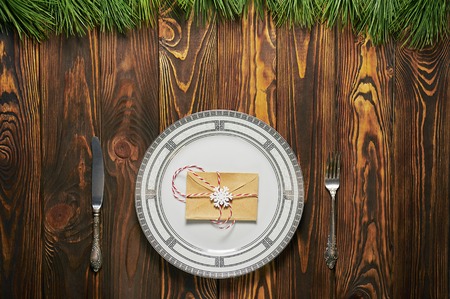 christmas table setting at brown wooden background with white plate, silver cutlery and paper envelope. xmas concept. top viewの写真素材