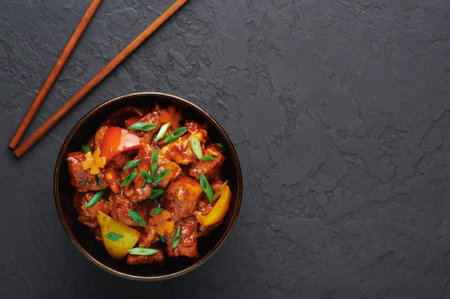 Chicken Manchurian in bowl at black concrete background. Chicken Manchurian is Indian Chinese cuisine dish with Chicken breasts, bell pepper, tomatoes, soy sauce. Copy spaceの写真素材